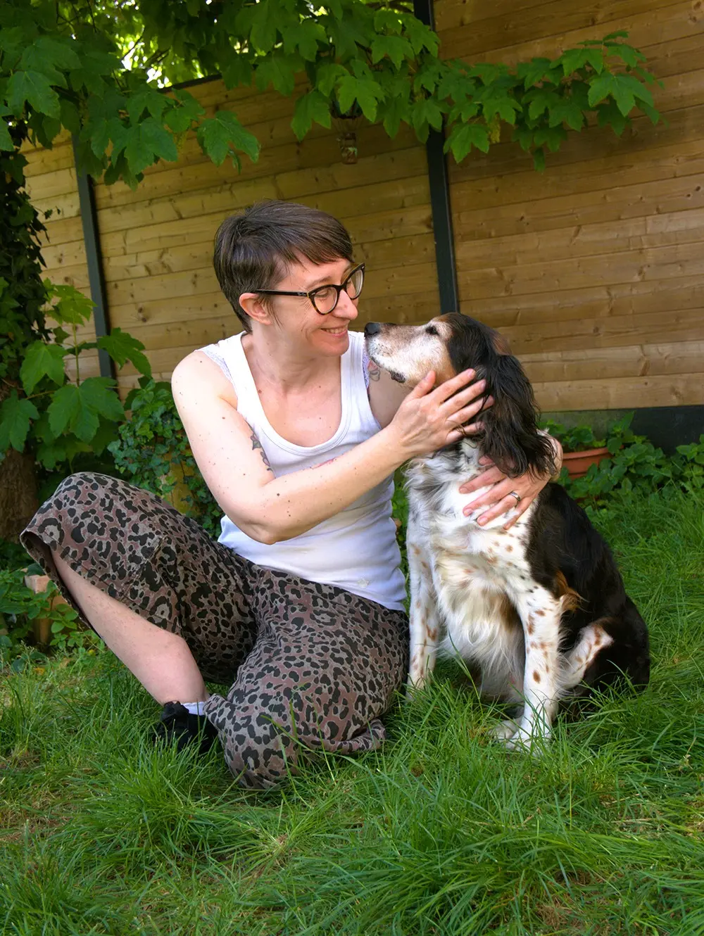 Morgane assise dans l'herbe, souriante, câlinant Guizmo, son chien croisé épagneul breton, devant une palissade en bois entourée de végétation.